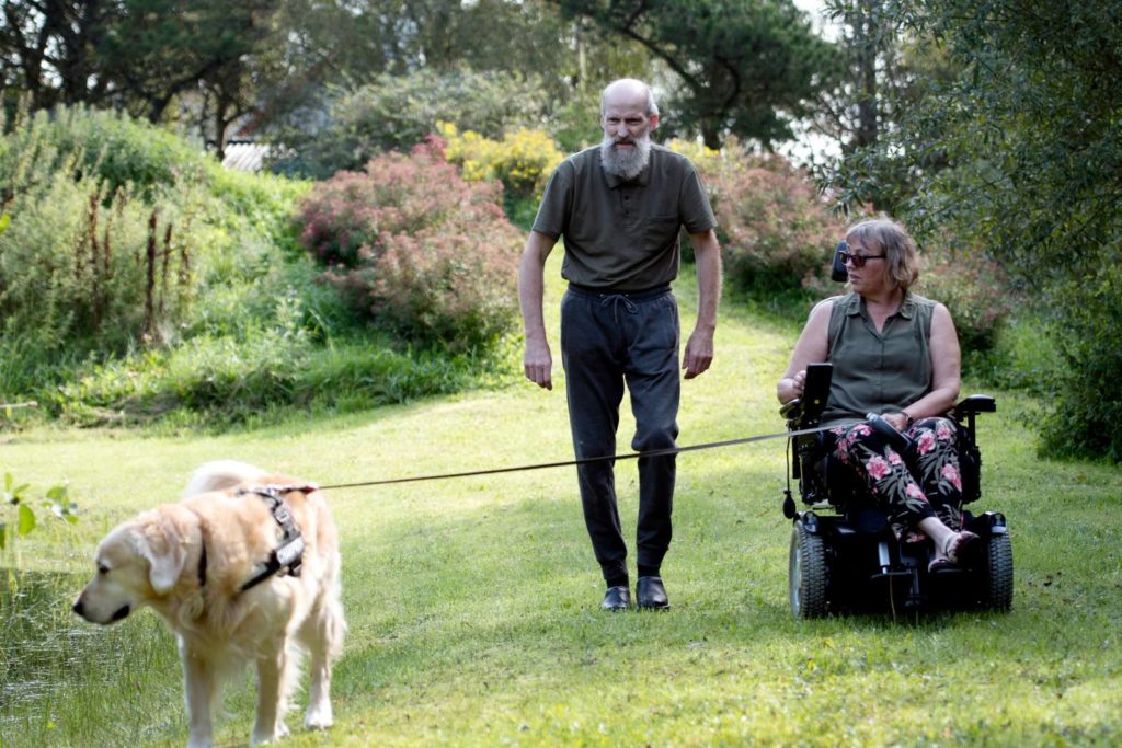 Kvinde i kørestol med hund i snor og en mand gående ved siden af.