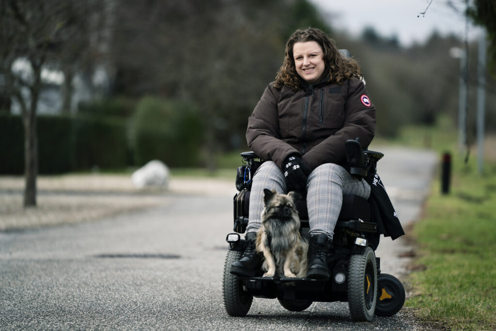 Katrine Jensen med sin hund Bailey siddende på fodstøtterne på kørestolen.